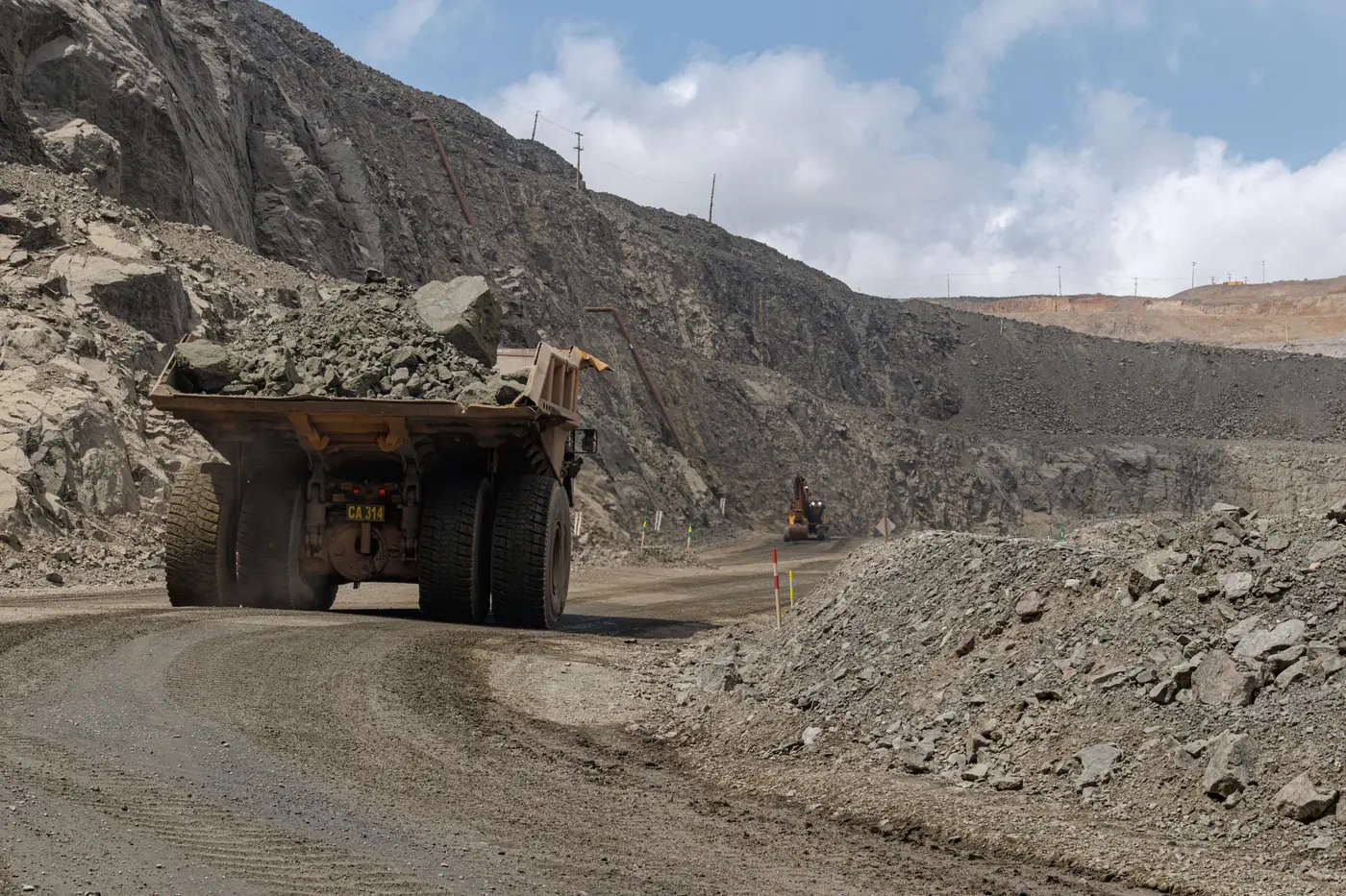 A loaded mining haul truck kicks up dust while driving down an unpaved open-pit road flanked by rock walls.